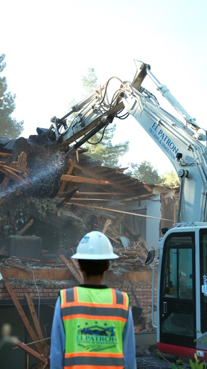 Excavator demolishing a house, worker watching